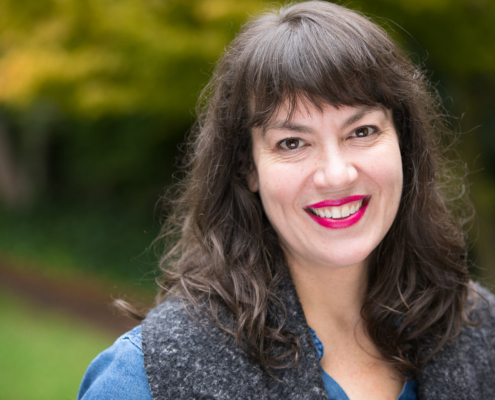 Portrait of a person smiling at the camera. Shoulder-length brown wavy hair with bangs, fair-skin, wearing a blue jacket