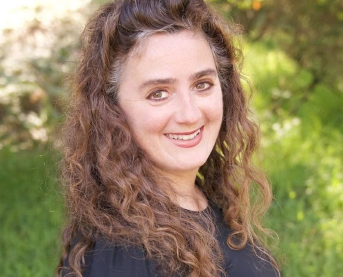 Portrait of a person smiling, in three-quarter view, looking into the camera. Long curly brown hair, fair-skinned, wearing a black shirt.