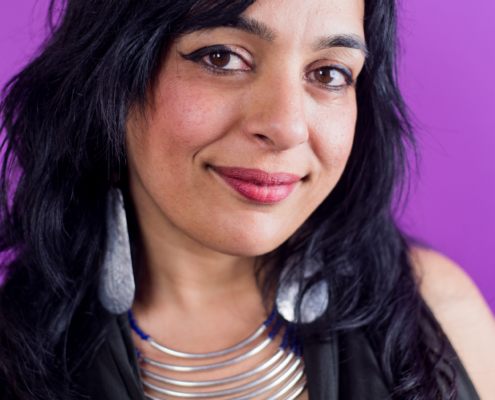 Portrait of a person looking at camera smiling, shoulder-length black hair and light brown skin, wearing large shell drop earrings, a multi-strand metal necklace, and black vest in front of a magenta background