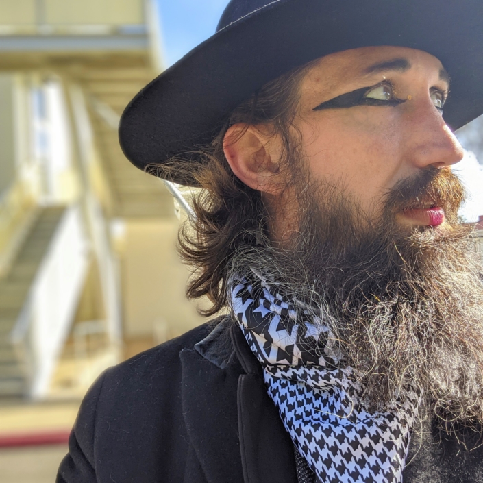 Portrait of Jason long looking to the right, brown hair and beard, wearing hat, stylized eyeliner, patterned scarf, and black jacket