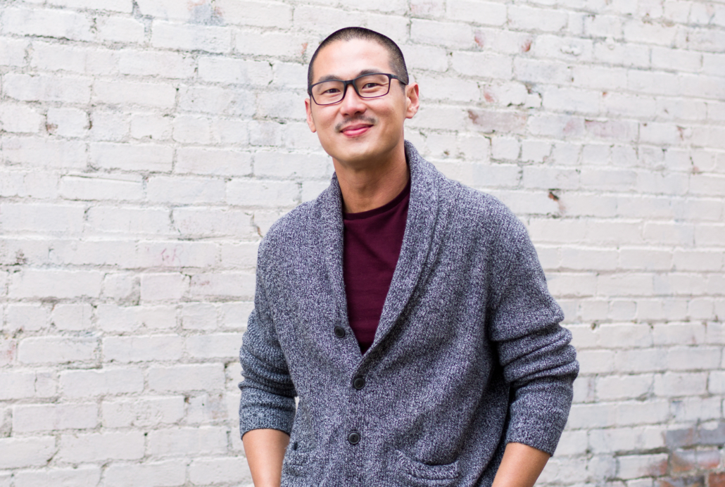 Edward Gunawan in a gray sweater and Burgundy T-shirtstanding before a whitewashed brick wall