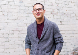 Edward Gunawan in a gray sweater and Burgundy T-shirtstanding before a whitewashed brick wall