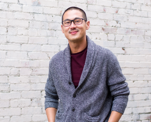 Edward Gunawan in a gray sweater and Burgundy T-shirtstanding before a whitewashed brick wall