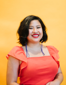 Michell Lin wearing a jeweled nose ring, long purple earrings, and an orange blouse in front of a bright yellow background