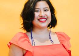 Michell Lin wearing a jeweled nose ring, long purple earrings, and an orange blouse in front of a bright yellow background