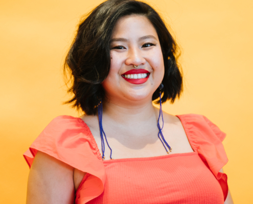 Michell Lin wearing a jeweled nose ring, long purple earrings, and an orange blouse in front of a bright yellow background