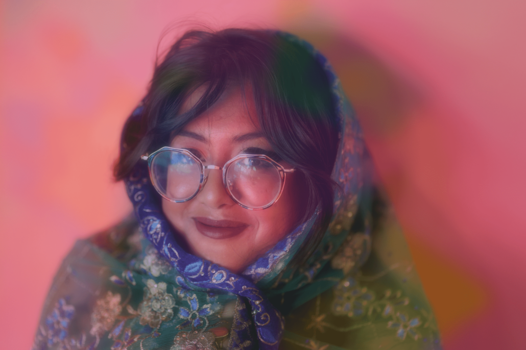 Sharmi Basu wrapped in a colorful scarf wearing wide circle glasses against a pink background