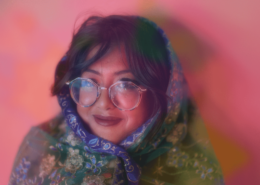Sharmi Basu wrapped in a colorful scarf wearing wide circle glasses against a pink background