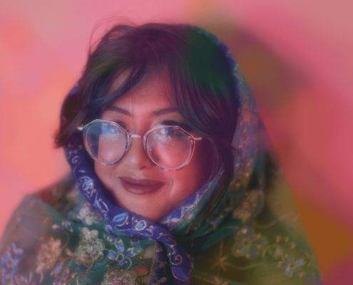 Sharmi Basu wrapped in a colorful scarf wearing wide circle glasses against a pink background